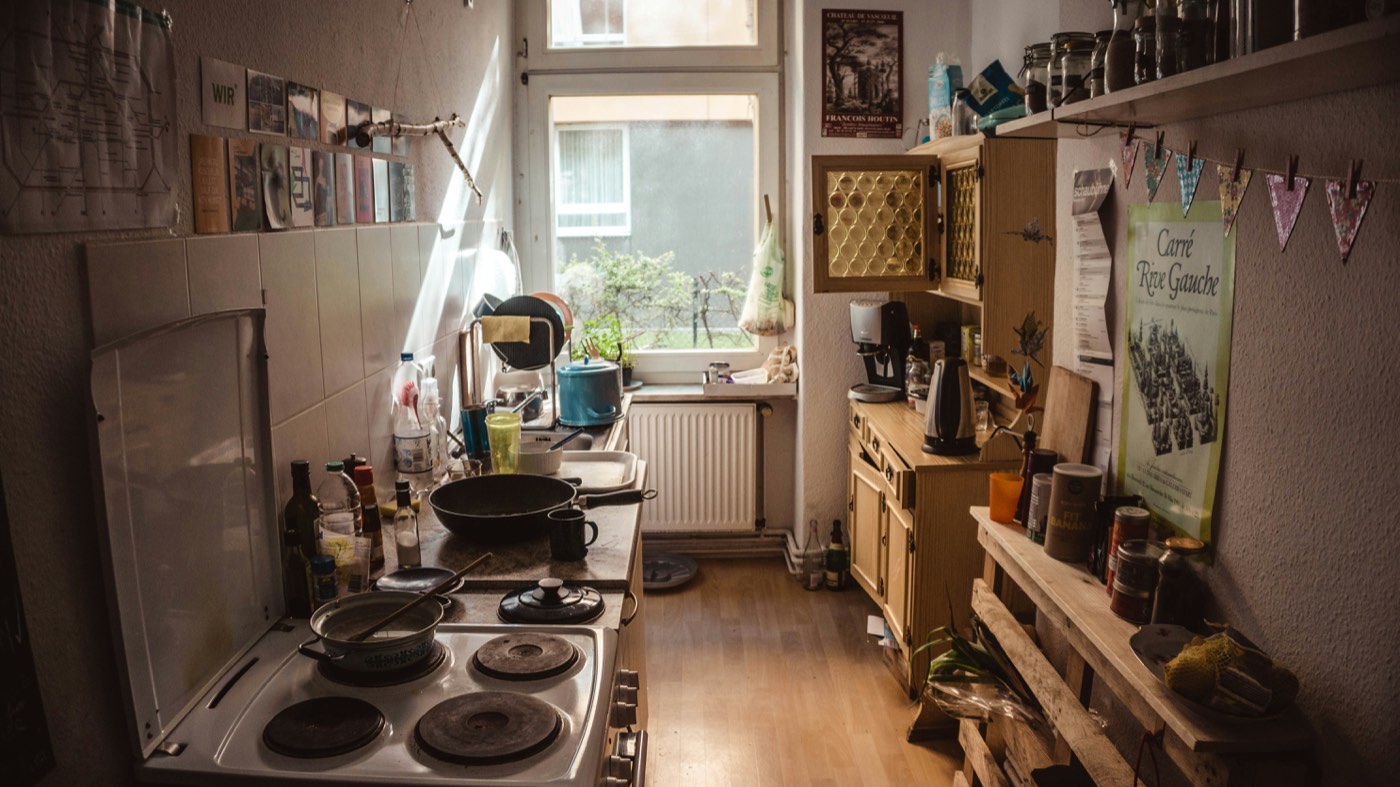 A small home kitchen with pans on the stove, postcards taped to the wall, and jars on a shelf by the window.