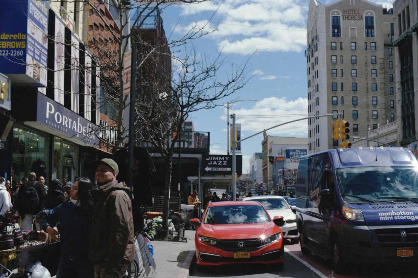 Two people stand on a sidewalk in front of a menswear shop, with street vendors, traffic, and Hotel Theresa in the background.