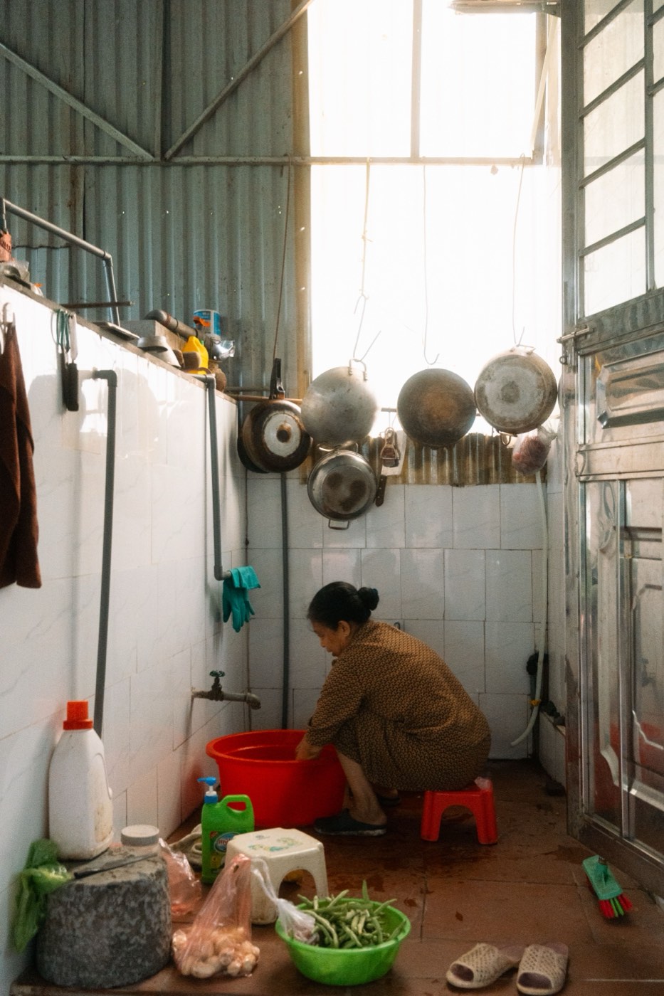 Someone washing vegetables in a small home kitchen, pots hanging on the wall, a plastic stool and sandals on the floor.