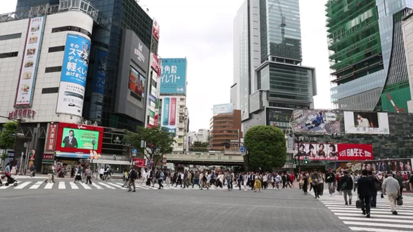 A large city crossing seen from a distance, crowds moving under grey sky.
